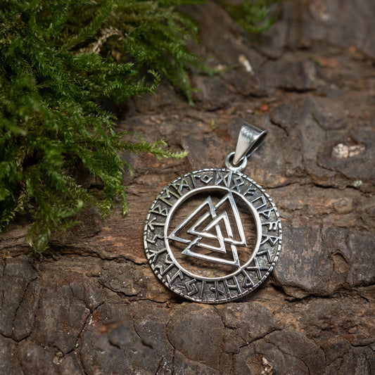 En Valknut Futhark Vedhæng 925s Sterling Sølv hviler på grov bark ved siden af en tot grøn mos. Det indviklede design og teksturerne står i kontrast til den robuste bark og bløde mos, hvilket fremkalder mystikken i nordisk mytologi.