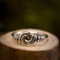 Indlæs billede i Gallery viewer, Sølvring Spiral 925s Sølv er elegant placeret på en træoverflade. Denne sølvring kan prale af et slående spiraldesign i midten, med indviklet wirework, der fremhæver spiralen, der afspejler vikingernes håndværk. Baggrunden er blødt sløret med jordnære og grønne nuancer.
