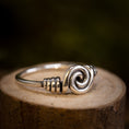 Indlæs billede i Gallery viewer, Et nærbillede af Sølvring Spiral 925s Sølv-ringen med et indviklet oprullet tråddesign i midten, der minder om Vikingerne håndværk. Ringen er placeret på en træoverflade med en mørk, sløret baggrund, der fremhæver ringens håndværk og skinnende, reflekterende egenskaber.
