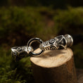 Indlæs billede i Gallery viewer, Et nærbillede af et Armbånd Fenris Ulven Glat Sølv-armbånd med to indviklet designede Fenrisulv-hoveder, der mødes ved en central ring. Armbåndet er placeret på en træstub, med en sløret naturlig grøn baggrund. Ulvehovederne udviser detaljerede graveringer og sorte accenter på deres træk.
