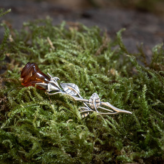 Rosebrosch med rav i 925s silver, formad som en stilk med blad, med äkta rav som liknar en blomknopp på grön mossa. Hantverket efterliknar en naturlig ros.