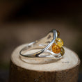 Indlæs billede i Gallery viewer, Et nærbillede af Sølvring Amber Drops 925s Sølv, en smukt udformet sølvring med et kunstnerisk, flydende design med tre runde ravsten i gule, orange og grønne nuancer. Ringen vises oven på en lille træstub, der fremhæver dens detaljerede håndværk mod en blødt sløret baggrund.
