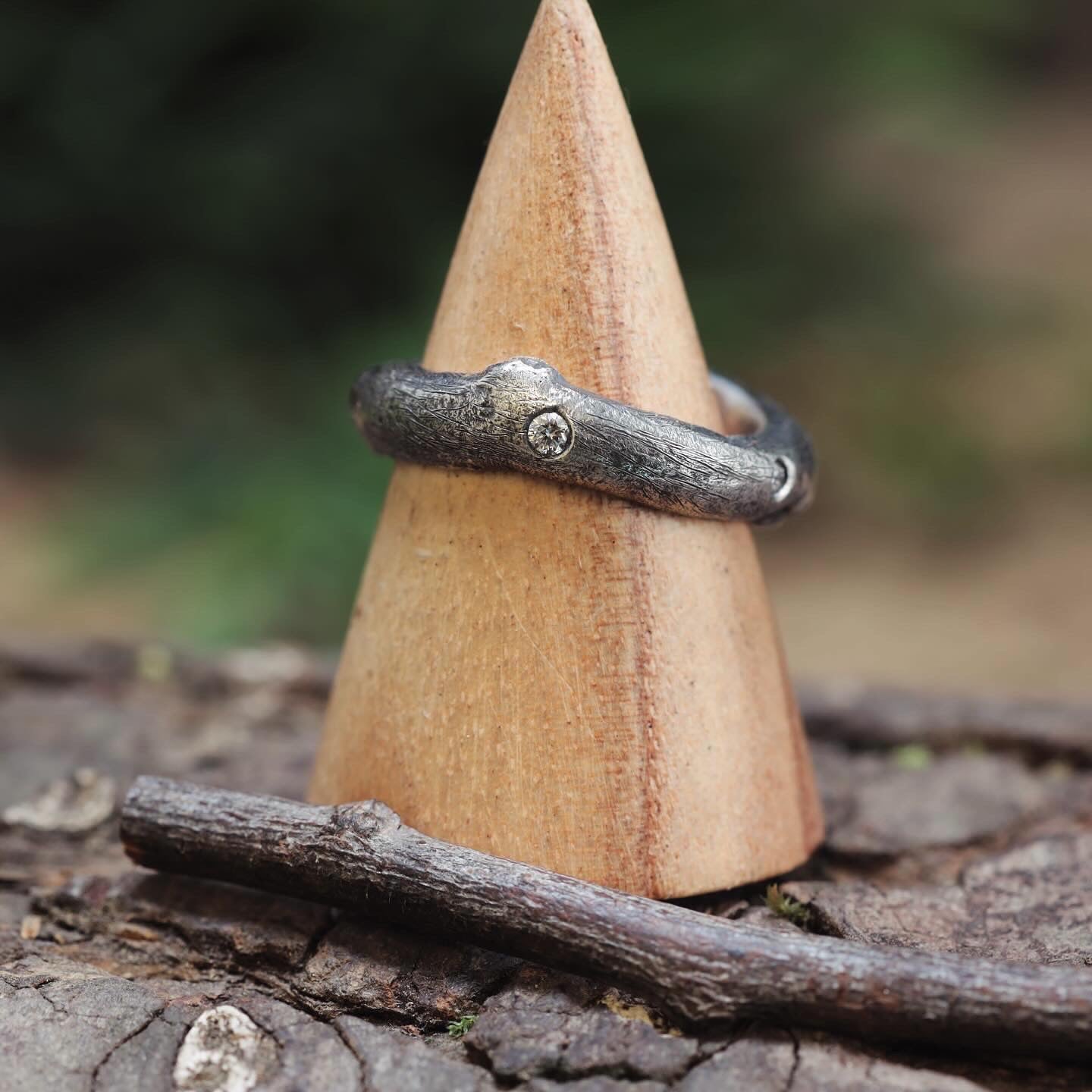 "Fingerring - Eryn Design - Branches", en sølvring, der viser et tekstureret, naturligt design med en lille indlejret ædelsten, vises på en trækegle. I baggrunden tilføjer bark tekstur, mens en kvist i forgrunden fuldender det rustikke æstetiske, indbegrebet af elegant dansk design.