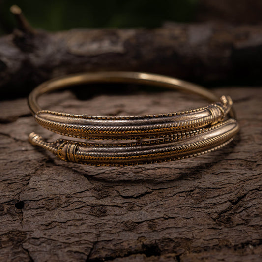 Et nærbillede af det omhyggeligt designede Armbånd Armbånd Midgårdsormen Bronze, med et strejf af Vikingetiden-design, hvilende på ru bark. Armringen viser detaljerede, fine mønstre og teksturer, der fremhæver dygtigt håndværk. Baggrunden er sløret, hvilket gør opmærksom på armringens udsøgte detaljer.