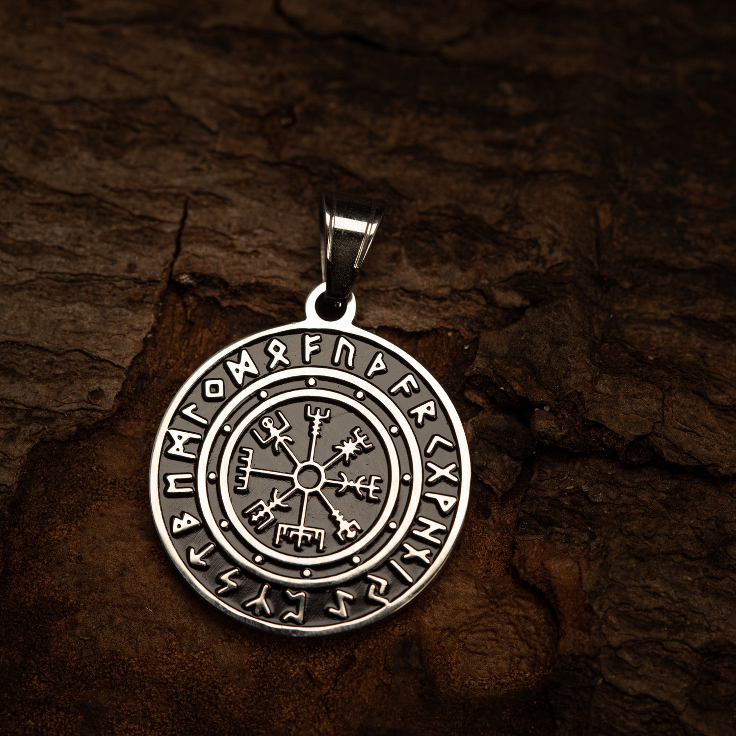 En cirkulær Rune Amulet Vedhæng Stål med nordiske runer og symboler, inklusive Vegvisir (et nordisk kompas) i midten. Stålvedhænget er placeret på en ru, brun baggrund, der ligner gammelt træ eller læder, med indviklede runeindskrifter i yderkanten.