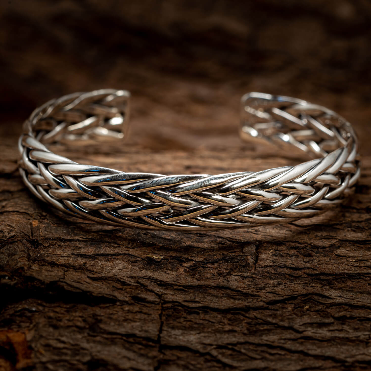 Et nærbillede af et indviklet vævet Armbånd Bangle Flettet 925s Sterling Sølv hvilende på en tekstureret træoverflade. Armbåndets polerede metaltråde er sammenflettet, hvilket skaber et komplekst, flettet design. Fokus fremhæver håndværket og detaljerne i dette udsøgte stykke.