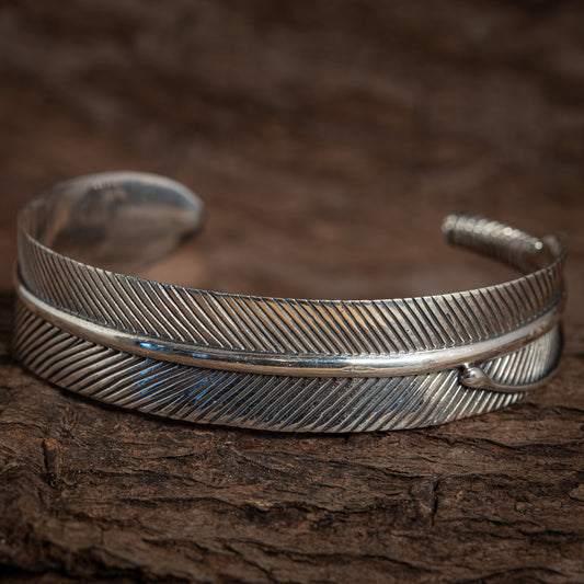 Et nærbillede af et indviklet udformet sølvmanchetarmbånd. Armbånd Bangle Feather 925s Sterling Sølv har detaljerede, ætsede diagonale linjer, der efterligner teksturen af en kragefjer med en poleret finish. Armbåndet er lavet af 925s Sterling Sølv og er sat mod en rustik, brun træbaggrund.