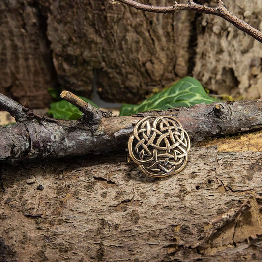 En smukt udformet, cirkulær Broche Keltisk knude Stor Bronze hviler på en ru trægren med grønne blade i baggrunden. Det indviklede evighedsknude-design skiller sig ud mod de naturlige, jordnære omgivelser af bark og kviste.