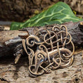 Indlæs billede i Gallery viewer, Broche Urnesstil Bronze, prydet med indviklede keltiske knudedesigns med stiliserede dyrehoveder, hviler på et stykke træbark. Denne autentiske bronzebroche fremkalder ånden fra Tröllaskógur-øen med grønne blade i baggrunden, der forstærker dens tidløse skønhed.
