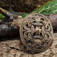 Indlæs billede i Gallery viewer, Broche Borrestil Bronze, en kompliceret designet broche i middelalderstil, der minder om autentisk vikingedesign, hviler på et stykke forvitret træ. Brochen har et udsmykket, symmetrisk mønster med sammenflettede detaljer og en central hævet knude, sat mod en rustik baggrund med grønt løv, der fanger essensen af Fæsted skatten.
