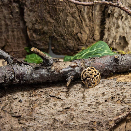 Broche Keltisk knude Bronze, et lille og indviklet udformet vedhæng med et keltisk knudedesign med antydninger af vikingemønster, er beliggende på en trægren. Den teksturerede bark og grønne blade i baggrunden skaber en naturlig ramme.