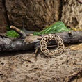 Indlæs billede i Gallery viewer, En Broche Keltisk knude Nål Bronze, der viser indviklede keltiske knudemønstre, er placeret på en rustik trægren. Grønne blade og stykker af bark omgiver brochen og forstærker de naturlige og jordnære omgivelser med dens autentiske bronzefinish, der minder om vikingetiden.
