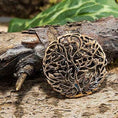 Indlæs billede i Gallery viewer, "Broche Yggdrasil Livets Træ Bronze", et indviklet udskåret rund bronzevedhæng, vises på en tekstureret træoverflade. Grøn vegetation i baggrunden tilføjer et naturligt præg til scenen.
