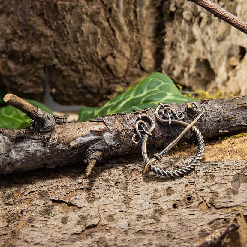 En Broche Ringnål Antik Bronze, prydet med indviklede løkker og et snoet ringdesign, der minder om Vikingetiden, er fastgjort til en forvitret trægren. Baggrunden har grønne blade og den teksturerede bark af et træ, hvilket skaber en naturlig, rustik indstilling.