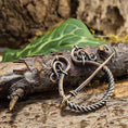 Indlæs billede i Gallery viewer, Et nærbillede af Broche Ringnål Antik Bronze, med indviklede løkker og et snoet design, der minder om traditionelle vikingebrocher, placeret på ru bark. I baggrunden skaber grønne blade og slørede naturlige elementer en udendørs rustik ramme.
