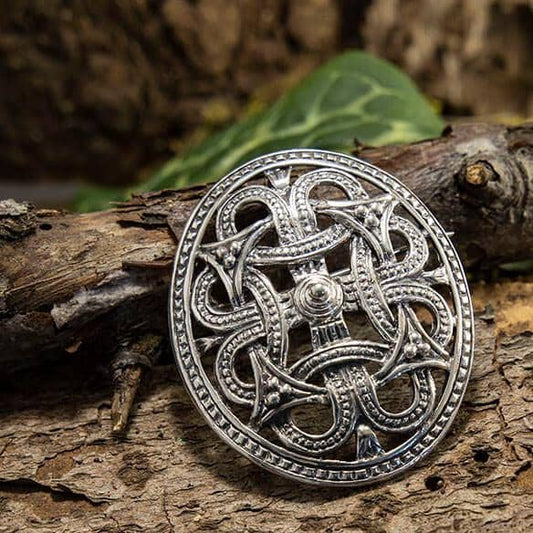 Broche Borrestil 925s Sterling Sølv, prydet med et delikat og komplekst keltisk knudedesign, er elegant udstillet på et stykke bark. Sammensætningen forstærkes af et grønt blad i baggrunden, der bidrager til den naturlige stemning. De omgivende rustikke omgivelser fremhæver brochens indviklede tekstur og overlegne håndværk, hvilket gør den til et lokkende stykke for enhver historieinteresseret.