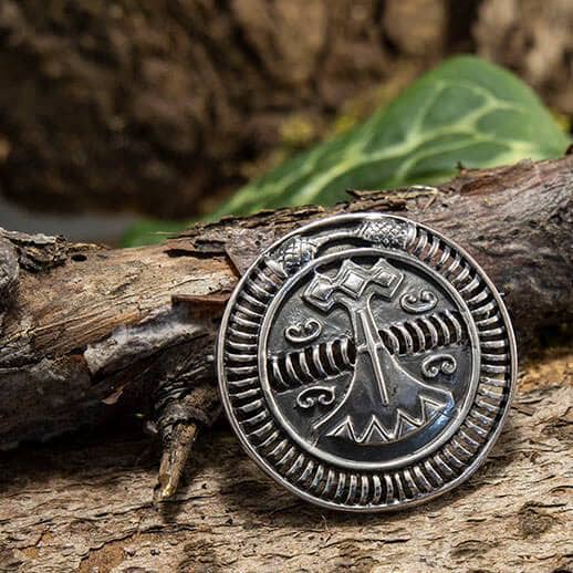 Et cirkulært vedhæng, der er indviklet designet med nordiske symboler, med et centralt billede af Thors hammer (Mjölnir) og lavet af ægte 925s Sterling Sølv. Vedhænget er placeret mod en rustik træbaggrund med grønne blade let synlige i den øverste del af billedet. Produktnavn: Broche Vikingeskib Spænde 925s Sterling Sølv