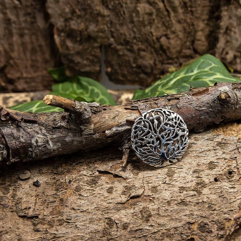 Broche Yggdrasil Livets Træ 925s Sterling Sølv, et cirkulært sølvtræ af livets vedhæng, der minder om Yggdrasil, hviler mod en tekstureret træstamme med bladgrønt i baggrunden. Dette indviklede design, der ligner Livets Træ, har sammenflettede grene og rødder, der symboliserer styrke og forbindelse til naturen.