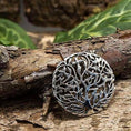 Indlæs billede i Gallery viewer, Broche Yggdrasil Livets Træ 925s Sterling Sølv, et rundt sølvvedhæng med et indviklet Yggdrasil-design, hviler på et stykke bark. Baggrunden har frodige grønne blade, der skaber en naturlig indstilling, der fremhæver vedhængets detaljerede håndværk.
