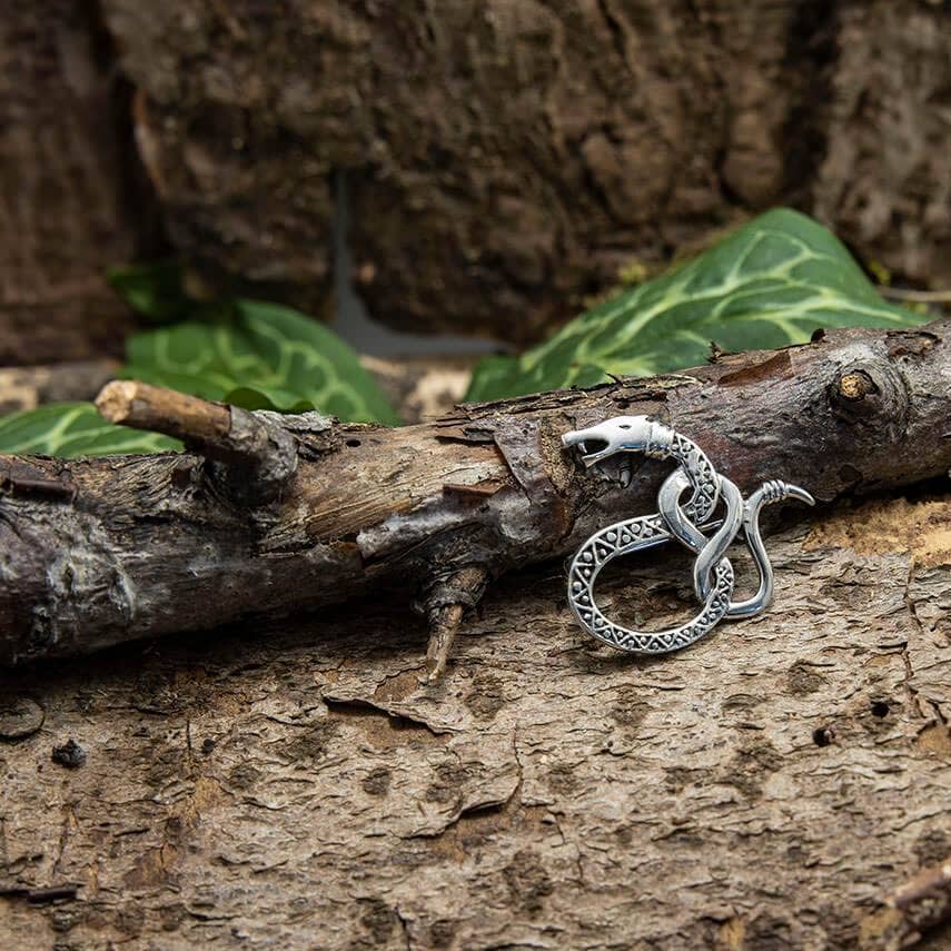 En sølv, indviklet designet Broche Slange Lille lavet af 925s Sterling Sølv hviler på en trægren sat mod en rustik baggrund med grønne blade. Brochen har en detaljeret slange viklet ind i en cirkel, der minder om design fra vikingetiden.