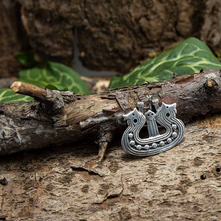 Et indviklet Broche Vikingeskib 925s Sterling Sølv, lavet af autentisk sølv, med tre dyrehoveder hviler på ru bark mod en naturlig baggrund af grønne blade. Denne autentiske vikingesmykke har detaljerede udskæringer og en række små cirkulære dekorationer langs dens nederste kant.