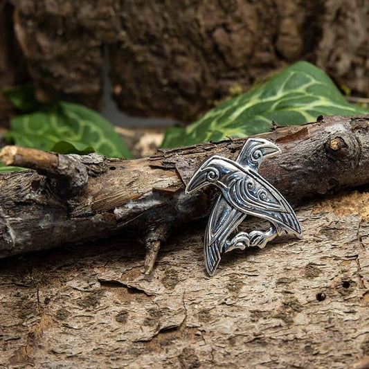 En Broche Hugin og Munin 925s Sterling Sølv, med to indviklede fugle, der muligvis repræsenterer Odins ravne, er fastgjort til et struktureret, rustikt stykke træ. Grønne blade er synlige i baggrunden, hvilket tilføjer en naturlig, jordnær indstilling til scenen.