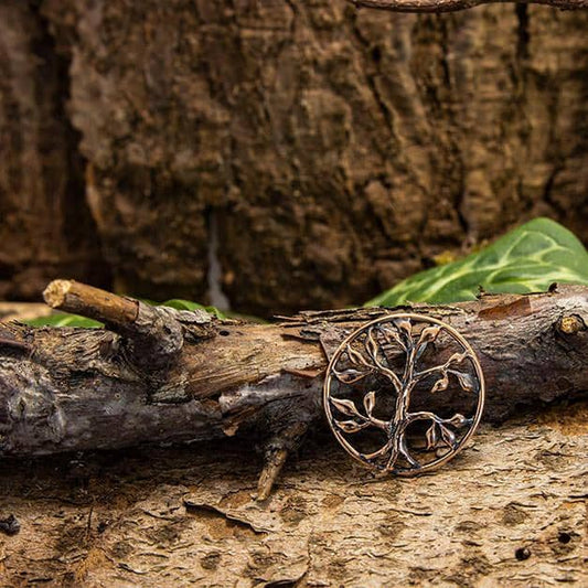 En Yggdrasil Livets Træ Blade Vedhæng Bronze, rund og indviklet designet, hviler på en ru, naturlig trægren. Baggrunden viser tekstureret bark og grønne blade, der forstærker den jordnære og rustikke æstetik knyttet til nordisk mytologi.