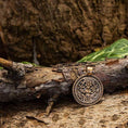 Indlæs billede i Gallery viewer, Amuletten Frigg Vedhæng Bronze, en rund medaljon med indviklede designs og symboler, hviler mod en ru, forvitret trægren på en skovbund. Baggrunden inkluderer grønne blade og træets teksturerede bark, hvilket forstærker den rustikke, naturlige scene.
