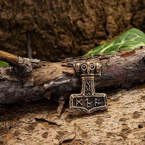 Thors Hammer Runer Vedhæng Bronze ligger på et stykke træ, indviklet designet med nordiske runer og en stiliseret hammerform, der symboliserer Thors Hammer fra nordisk mytologi. Vedhængen er lavet i bronze og er sat på baggrund af bark og grønne blade.