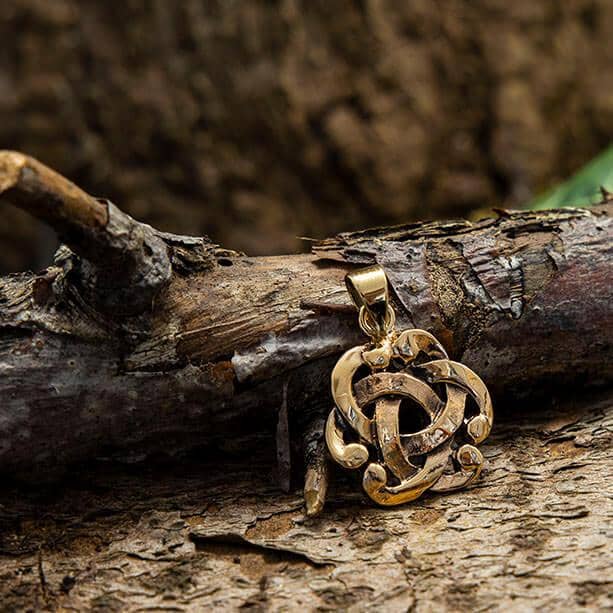 Et bronzevedhæng ved navn Wayland knuden Vedhæng, med et indviklet, cirkulært Keltisk knude-design, hviler på en ru trægren placeret på jorden med en brun, naturlig baggrund.