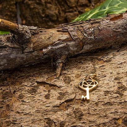 Nøglen til Himmeriget Mini Vedhæng Bronze-vedhæng, formet som en nøgle, hviler på den ru bark af et træ og blander sig problemfrit med jordfarverne. En gren og nogle grønne blade er synlige i baggrunden, hvilket forstærker de naturlige omgivelser.