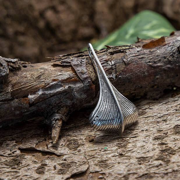 Et Gokstad skibet Vedhæng 925s Sølv vedhæng, formet som et udsmykket blad, hviler på et groft stykke bark. Baggrunden har en jordagtig tekstur med slørede grønne blade. Vedhænget er lavet af 925s Sterling Sølv og har detaljerede linjer og en glat, metallisk finish, der minder om Vikingeskibsvedhæng fundet på Gokstadskibet.