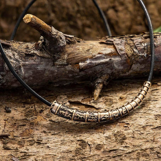 Nærbillede af Guldhorn Vedhæng Bronze med Læderkæde mod rustik træbaggrund. Halskæden har en sort lædersnor med et indviklet detaljeret ornamentalt bronzesegment i midten, prydet med forskellige indgraverede symboler og mønstre, der minder om Vikingetidens artefakter. Baggrunden inkluderer en ru trægren.