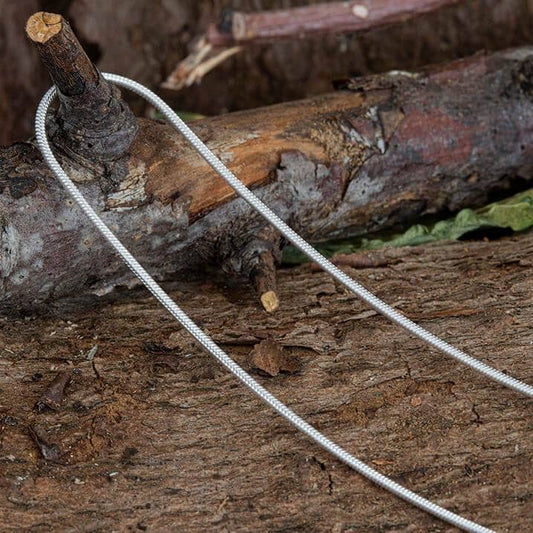 En metallsågkabel ligger på en grov träyta, troligen för utomhus- eller överlevnadsbruk. Den är hoprullad med två öglor som handtag. Bland bark och löv skymtar en halskedja ormkedja 925s silver 2 mm med oväntad elegans.