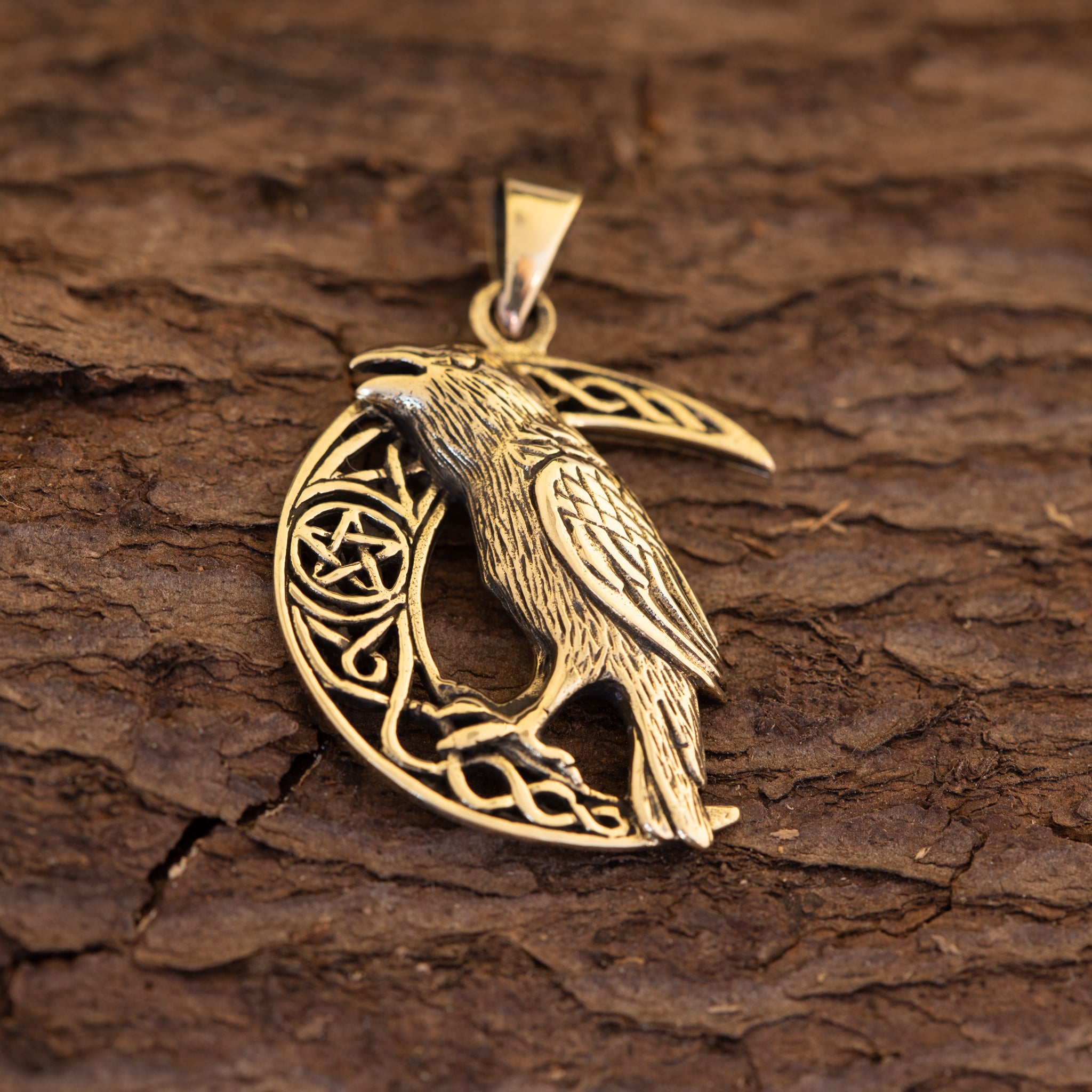 Vedhæng Crow Moon Bronze, et vedhæng med en ravn anbragt på en halvmåne og indgraveret med indviklede keltiske designs, hviler på en tekstureret træoverflade.