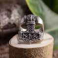 Indlæs billede i Gallery viewer, Sølvring Thors Hammer 925s Sølv, en ring i sterling sølv med indviklede nordiske designs, sidder på en træsokkel. Thors-hammeren fungerer som midtpunktet, prydet med runer og detaljerede udskæringer. Dette udsøgte stykke, fås i forskellige ringstørrelser, er sat mod grønt løv og en sløret naturlig indstilling.
