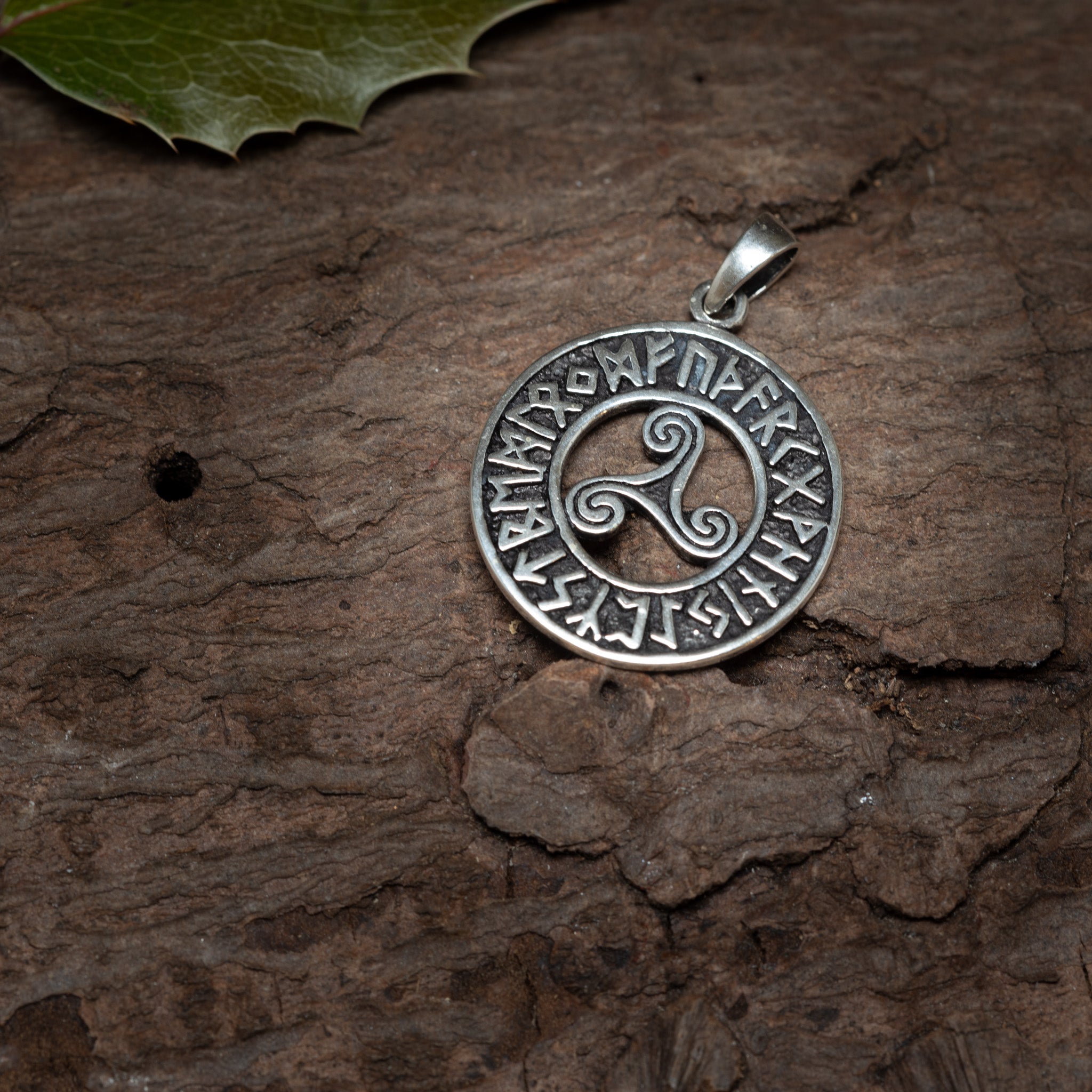 Vedhæng Tre Flige m. Runer 925s Sølv vedhæng viser et triskelion-symbol i midten, omkranset af runelignende tegn fra futhark-runealfabetet indskrevet rundt om den ydre kant. 925s Sterling Silver vedhænget hviler på en tekstureret træoverflade med et grønt kristtornblad synligt i øverste venstre hjørne.