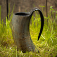 Indlæs billede i Gallery viewer, Vikingekrus Nature Horn (600-800 ml), et rustikt, buet drikkehorn med en poleret finish og fremtrædende håndtag, står på en plet af grønt græs. Træbaggrunden og de naturlige omgivelser forstærker dens middelalderlige, historiske tiltrækningskraft, der minder om den ældgamle vikingekrus, der blev brugt i oldtiden.
