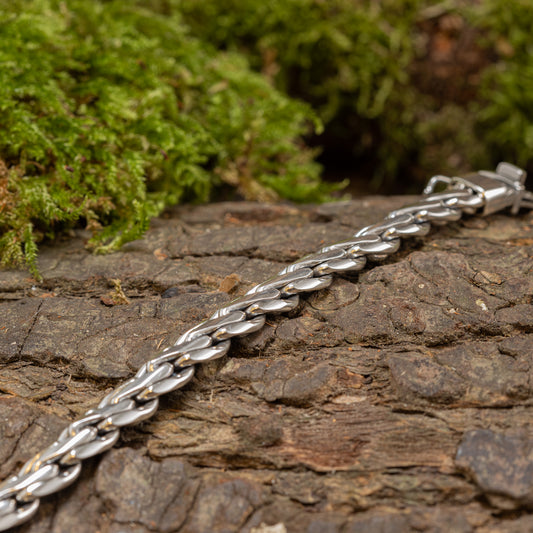 Et Armbånd ByKila Grov Flet 5,2mm 925s Sølv armbånd ligger på en ru træbark overflade med grønt mos i baggrunden. Armbåndet har sammenlåsende led og en kasselåslås, der skaber en scene, der emmer af rustikke, naturinspirerede omgivelser.