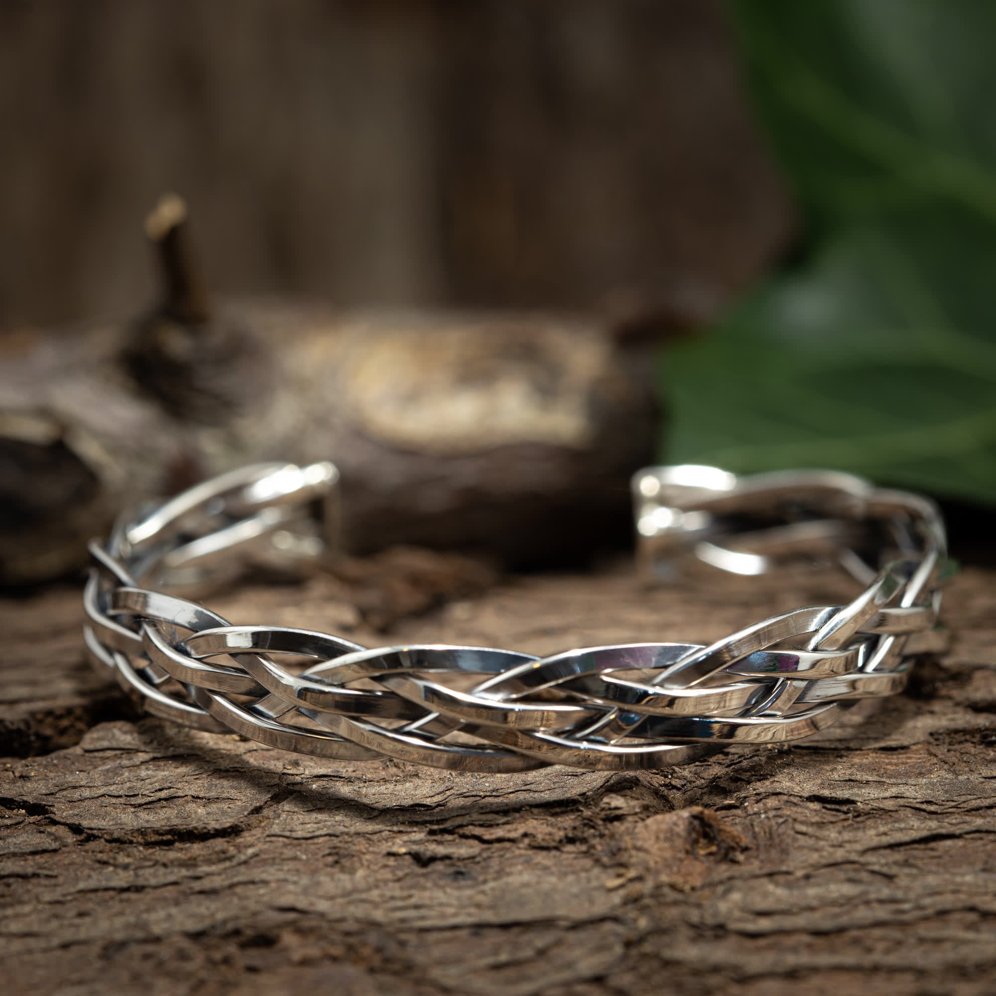 Et sølv Armbånd Bangle Flet 925s Sterling Sølv armbånd, lavet af 925s Sterling sølv, hviler på en ru, brun træoverflade med slørede grene og blade i baggrunden. Det indviklede vævet armbånd har et smukt sammenflettet mønster.