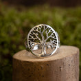 Indlæs billede i Gallery viewer, Livets Træ Yggdrasil Fingerring 925s Sterling Sølv viser et indviklet trædesign med bare grene indkapslet i en cirkulær ramme. Dette detaljerede stykke er elegant vist på et lille stykke træ mod en sløret grøn baggrund.
