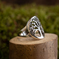 Indlæs billede i Gallery viewer, En Livets Træ Yggdrasil Fingerring 925s Sterling Sølv, med et detaljeret træmotiv, vises på en cylindrisk stensokkel. Baggrunden er sløret med grønne nuancer, hvilket tyder på løv eller udendørs landskaber.
