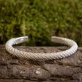 Indlæs billede i Gallery viewer, Et Armbånd Bangle Empor 925s Sterling Sølv hviler på et stykke bark, med grønt mos sløret i baggrunden. Armbåndet har et indviklet, vævet design.
