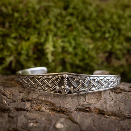 Et Armbånd Armbånd Mjølnir i 925'erne Sterling Sølv, med indviklede keltiske knudedesigns og et centralt Thors hammersymbol, vises på en træoverflade med en sløret grøn, mosbeklædt baggrund. Det detaljerede håndværk og mønstret er tydeligt synligt.