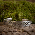 Indlæs billede i Gallery viewer, Armbånd Bangle Triskele 925s Sterling Sølv-armbånd hviler på en struktureret overflade og viser indviklede keltiske knudedesigns og et centralt cirkulært triskele-motiv. Baggrunden af sløret grønt understreger armbåndets detaljerede håndværk.
