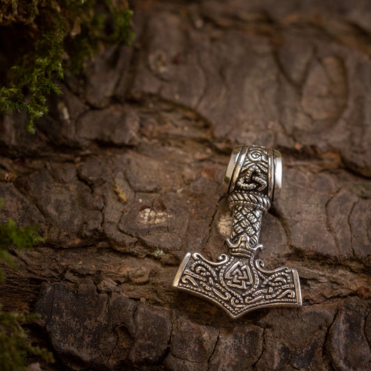 Thors Hammer Vedhæng 925s Sterling sølv Celtic, et sølvvedhæng designet til at ligne Mjolnir (Thors hammer), har indviklede nordiske og keltiske mønstre. Den fint udformede 925 sterling sølv hammer vises mod en struktureret træbark baggrund, med mos rundt om kanterne, der tilføjer et naturligt præg til billedet.