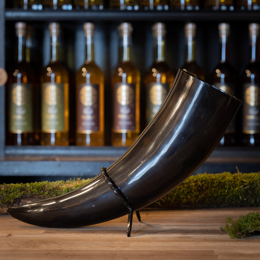 En polerad svart Drickshorn Giant 1,5 liter står på ett stativ framför en hylla med flaskor av olika sprit, alla med guldprydda cirkel logotyper. Mossa på träytan ger displayen en rustik vikingamarknadskänsla.