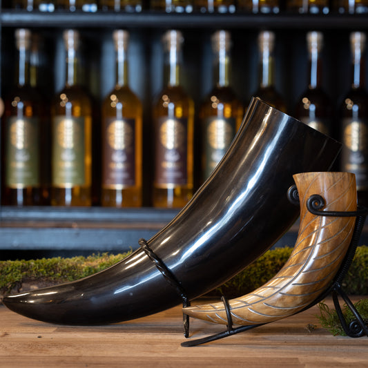Två utsökt utformade Drickshorn, inklusive Drickshorn Giant 1,5 liter, i olika storlekar och färger visas på ett träbord. Varje buffelhorn står på ett metallstativ, med flaskor i bakgrunden på hyllor. Miljön är rustik och elegant, som traditionella vikingamarknader.