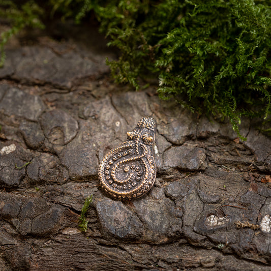 Vedhæng Slangen fra Gørding Bronze, en detaljeret metallisk amulet med et indviklet firbendesign, sidder på bark med ru tekstur, omgivet af grønt mos.