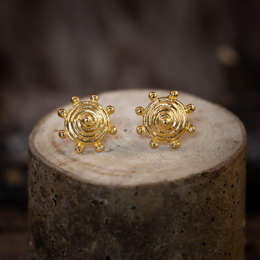 Närbild av örhängen Spiral Sköld, förgyllda 925s 925 Sterling Silver-örhängen, med ett centralt cirkulärt mönster av koncentriska ringar och små runda utskott längs kanten, på ett rustikt cylindriskt trästativ.